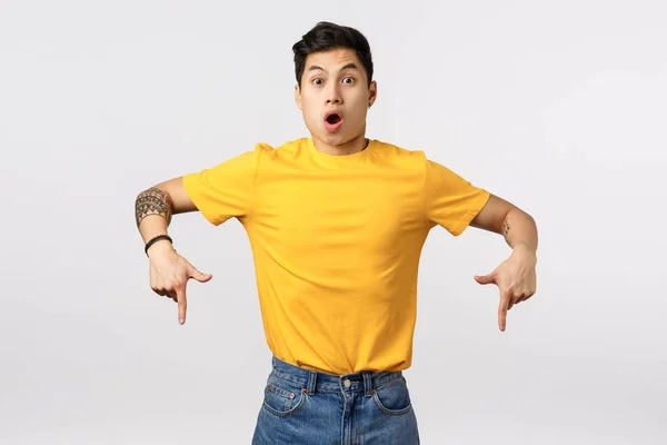 Les gars attention capturée sur la promo incroyable. Homme asiatique étonné et impressionné en t-shirt jaune, pointant vers le bas, lèvres pliantes étonnées, parlant de nouvelle offre promotionnelle, fond blanc debout excité — Photo