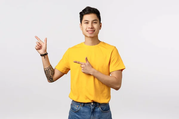 Atraktivní asijský muž moderní s potetovanou rukou, nosit žluté tričko, propagovat firemní banner, vánoční zimní prodej, usměvavý spokojen, doporučit použití produktu, stojící bílé pozadí reklama — Stock fotografie