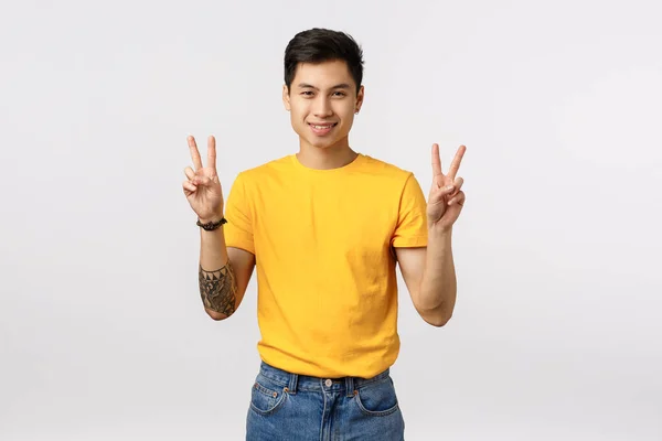 Elegante bem parecido asiático homem com tatuagens em camiseta amarela, mostrando sinais de paz, gesto de vitória, sorrindo alegremente, enviar vibrações positivas, se divertindo, de pé em humor brincalhão, fundo branco — Fotografia de Stock