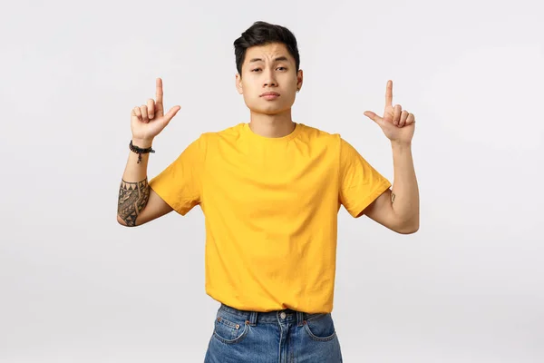 Skeptical, not impressed handsome asian man in yellow t-shirt, pointing up, frowning and grimace with dislike, unbothered standing white background in yellow t-shirt, unimpressed with lame offer — 스톡 사진