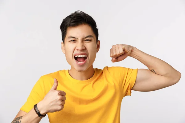 Primer plano tiro atrevido y seguro de buen aspecto asiático hombre en camiseta amarilla, mostrando el músculo y el pulgar hacia arriba, gritando animado y emocionado, sintiéndose optimista y entusiasta para comenzar a entrenar — Foto de Stock