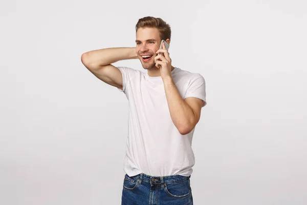 Happy flattered and pleased handsome blond guy receive praises or good news via phone call, holding smartphone near ear, touching neck silly and embarassed, standing white background — Stock Photo, Image