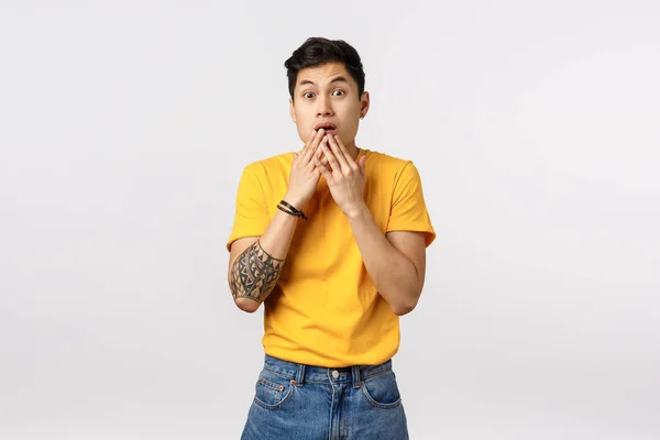 Scared, shocked young astonished asian guy in yellow t-shirt, gasping, cover opened mouth with palms and stare camera impressed, gossiping, realise something shocking, stand white background — Stock Photo, Image