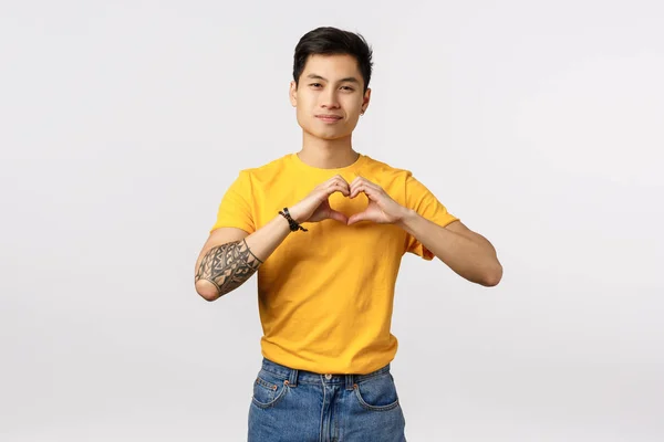 O amor está no seu coração. bonito e bonito jovem ásia tatuado hipster cara no amarelo t-shirt expressar afeto fazendo relacionamento símbolo sobre peito e sorrindo com Quente sorriso, branco fundo — Fotografia de Stock