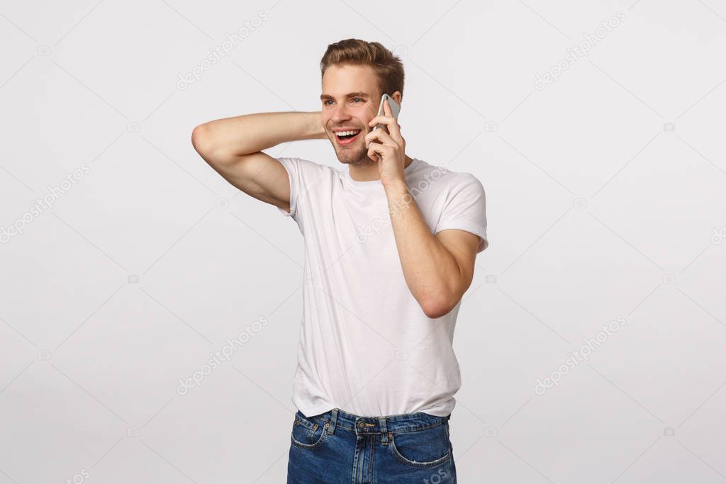 Happy flattered and pleased handsome blond guy receive praises or good news via phone call, holding smartphone near ear, touching neck silly and embarassed, standing white background