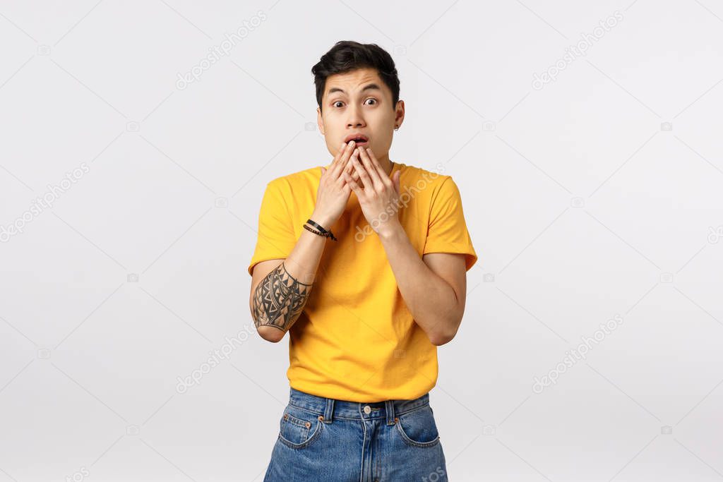 Scared, shocked young astonished asian guy in yellow t-shirt, gasping, cover opened mouth with palms and stare camera impressed, gossiping, realise something shocking, stand white background