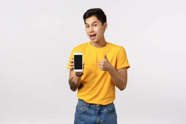 Atraente asiático cara em amarelo elegante t-shirt com tatuagens, mostrando polegar para cima e exibição de smartphone como oferecendo o download do cliente incrível aplicativo, aplicativo de edição, jogo ou página corporativa — Fotografia de Stock