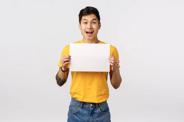 Entusiastiska, glada stilig kinesisk kille med tatueringar, hålla skylt, tomt papper över bröstet, främja företagets erbjudande, ler med glädje och lycka, stående vit bakgrund annonsera — Stockfoto