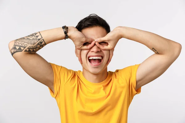 Concepto de diversión, emoción y entusiasmo. Alegre y despreocupado juguetón asiático tatuado chico en camiseta amarilla, haciendo gafas o máscara de superhéroe con los dedos sobre los ojos, de pie fondo blanco alegre —  Fotos de Stock