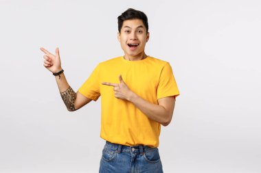 Heyecanlı, mutlu Asyalı genç adam, sarı tişört, kot pantolon giy, şaşkın bir şekilde gülümse, kış satışını tavsiye etmekten büyülen, indirim yap, beyaz arka planda dur.