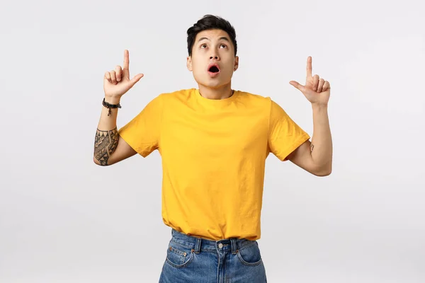 Wow, omg look at that. Astonished, impressed asian cute male model in yellow t-shirt, with tattoos, stare and pointing up with dropped jaw, speechless standing white background, fascinated — Stock Photo, Image