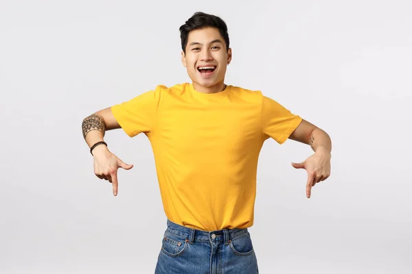 Bezstarostný, radostný okouzlující mladý asijský milennial chlap ve žlutém tričku, s tetováním, ukazující dolů ukazováčky a usmívající se fotoaparát, rada obrátit pozornost dolů promo, bílé pozadí — Stock fotografie