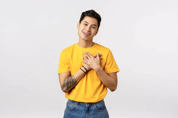 Lovely, touched asian guy with tattoos in yellow t-shirt, press hands to heart, sighing with affection or admiration, smiling thanking, grateful for gift, thankful stand white background — Stock Photo, Image