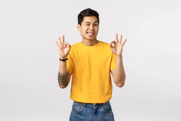 Relax, I got everything under control. Happy cheerful asian man in yellow t-shirt, wink and show OK, okay gesture as telling all good, approve product, like your choice, smiling and agree — Stock Photo, Image