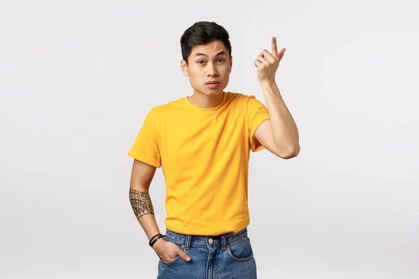 Wat is er aan de hand? Verveelde Aziatische jongeman in geel t-shirt, met tatoeages, wijsvinger opsteken in ontzetting en irritatie, wenkbrauwen opheffen als gelul, raar gesprek, witte achtergrond — Stockfoto