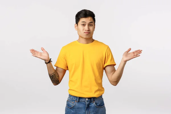 Sceptische en lastige jonge arrogante aziatische man met tatoeages, geel t-shirt dragen, handen zijwaarts spreiden in verbijstering en verwarring, besluiteloos geen antwoord geven, niets weten — Stockfoto