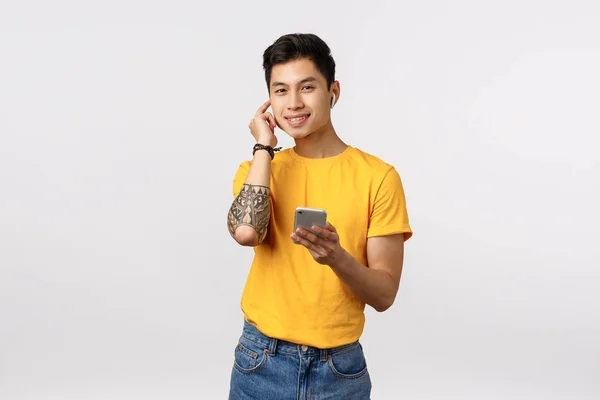 Handsome stylish chinese guy with tattoos, put on wireless headphones, holding smartphone and smiling pleased, listen music, enjoying nice earphones bit, standing white background