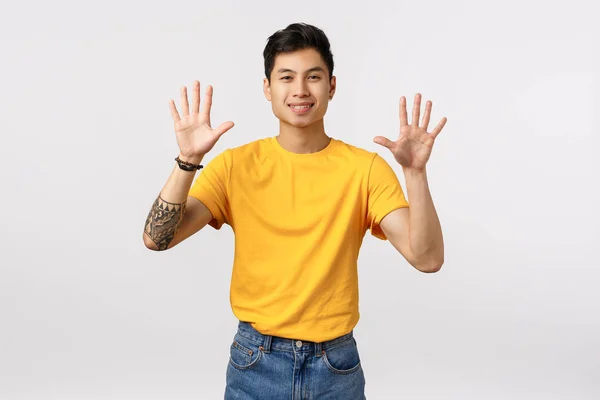 黄色のTシャツの陽気でフレンドリーな外観の魅力的なアジア人男性,入れ墨を持ちます, 10本の指を示します,腕を上げます,注文ダース,楽しそうに笑顔,白い背景に立ってアップビート — ストック写真