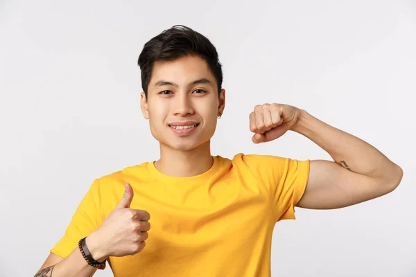 Tiden pumpar upp den. Attraktiv ung asiatisk man i gul t-shirt visar muskler och tumme-up, ler, uppmuntra göra fysiska övningar, komma gym tillsammans, vill få starka biceps — Stockfoto
