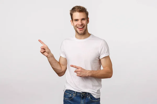 Hey, check this out, great price, cool product. Charming smiling blond male model with bristle, pointing upper left corner and curiously grinning as asking question, consult shop assistant — Stock Photo, Image