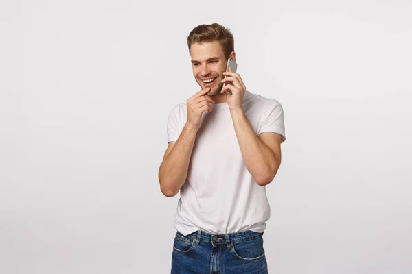 Intrigued and happy excited blond guy receive invitation party via phone call, holding smartphone near ear, talking on mobile, touching lip and smiling tempting, have conversation, white background — Stock Photo, Image