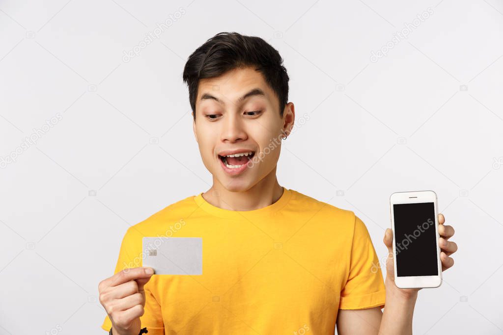 Close-up shot enthusiastic guy decided put money deposit, get credit to pay for trip, holding smartphone showing display and banking card, smiling amused, standing white background