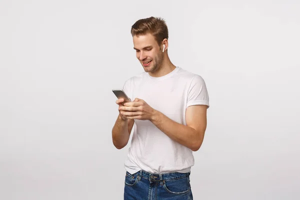 Χαμογελαστός χαρούμενος ξανθός Ευρωπαίος με λευκό t-shirt, τζιν, κρατώντας smartphone, φορούν ασύρματα ακουστικά, εμφάνιση mobile οθόνη, ανάγνωση μήνυμα, επιλέξτε τραγούδι, λευκό φόντο — Φωτογραφία Αρχείου