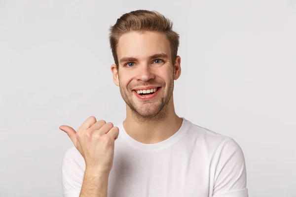 Close-up studio portrait blond handsome and joyful, smiling man asking friend opinion as pointing thumb left, discuss company product, grinning cheerful, standing white background — Stock Photo, Image