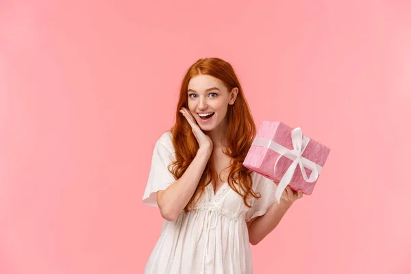 Sorprendido, tonta y encantadora chica pelirroja femenina recibir regalo, mirada divertida y se preguntó, no esperaba, sonriendo complacido, sosteniendo regalo envuelto, maravilla lo que está dentro, de pie fondo rosa — Foto de Stock