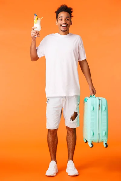 Disparo vertical de cuerpo entero tipo afro-americano feliz de vacaciones, sosteniendo el equipaje como viaje, diciendo hola amigos en el hotel, bebiendo cóctel y sonriendo felizmente. fondo naranja de pie optimista —  Fotos de Stock