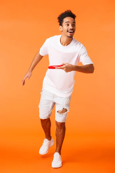 Full-length vertical tiro feliz, carismático afro-americano desportivo cara como jogos ativos ao ar livre, jogando frisbee em amigo e sorrindo, desfrutando de belo dia ensolarado de verão, fundo laranja — Fotografia de Stock