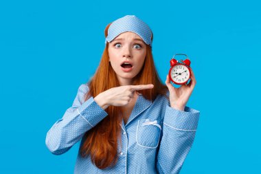 Şok olmuş ve endişeli, endişeli kızıl saçlı kız çalar saate sinirli bir ifadeyle bakıyor, geç kalıyor, doğru zamanı bilmiyor, gece elbisesiyle mavi arka planda duruyor.