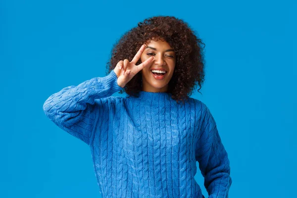 Gondtalan szép, karizmatikus afro-amerikai nő azt mondják, cheeze pózol örömmel és vidáman, mutatja béke jele felett kacsintás szem és mosoly, nevetve boldogan, élvezze a téli ünnepek, kék háttér — Stock Fotó