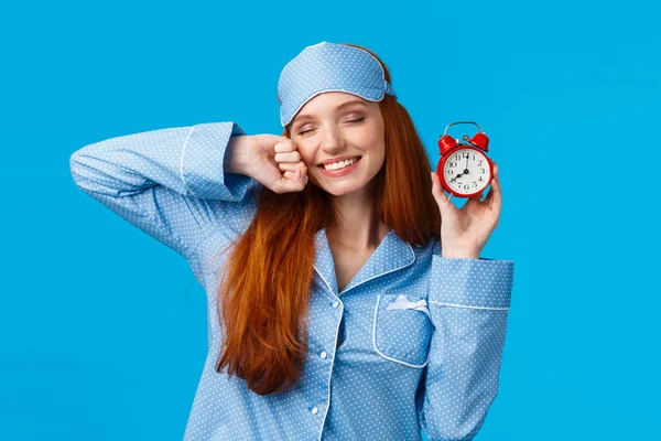 Girl slept well, wake up energized, sleepy stretching with closed eyes and lovely smile, holding red cute clock, set up alarm to be work in time, wearing pyjama and sleep mask, blue background