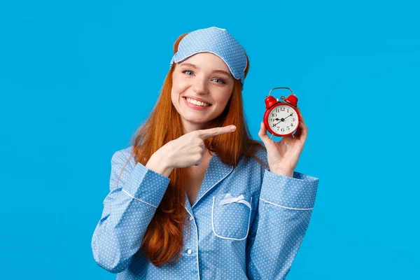 Не забудь проснуться вовремя. Веселая и милая расслабленная рыжая женщина в пижаме и маске для сна, держа красные часы, указывая на тревогу, как установлено перед сном, стоя синий фон — стоковое фото