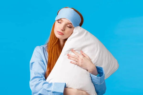 Beauty, female and lifestyle concept. Relaxed and relieved lovely redhead woman in sleep mask and pyjama, close eyes sleepy, having sweet dreams, hugging pillow, standing blue background — Stock Photo, Image