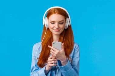 Belden yukarı portre kulaklıklı yakışıklı seksi üniversiteli kız, müzik dinliyor, sabahları haber yayını yapıyor, akıllı telefondan sosyal medyayı kontrol ediyor, pijamayla mavi arka planda duruyor.