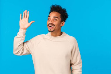 Hey merhaba, buradayım. Çekici dost canlısı Afro-Amerikan hippi erkek afro saç kesimi, bıyıklı, kalabalık bir arkadaş görüyor, sol üst köşeye bakıyor, el sallıyor selam veriyor, gülümsüyor, mavi arka plan