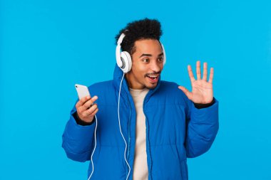 Teknoloji, sohbet konsepti. Şık, çekici, Afro-Amerikan erkek öğrenci, yastıklı kışlık ceketli, müzik dinleyen üniversite yolunda, akıllı telefon kulaklığı takan, el sallayan, selam veren,