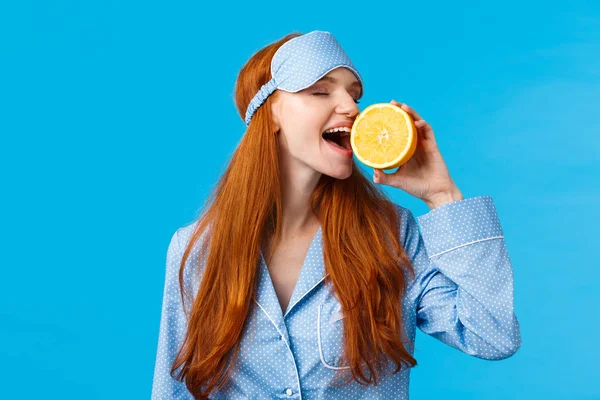 Delicious fruit starting morning healthy. Waist-up portrait attractive sleepy, pretty redhead girl in sleep mask and nightwear, biting slice of orange eating tasty, standing blue background