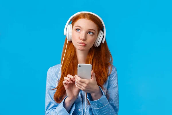 Przemyślana i kreatywna, zadumana, biała ruda kobieta, seksowna dziewczyna w nocnych ciuchach, patrząca w górę myśląca, nosząca słuchawki słuchająca muzyki lub podcastu, trzymająca smartfona, niebieskie tło — Zdjęcie stockowe