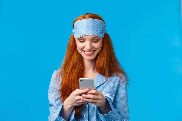 Mobile addication, technology and beauty concept. Cheerful girl waking up and grab phone, checking messages, scrolling news feed on smartphone wearing nightwear, sleep mask, smiling