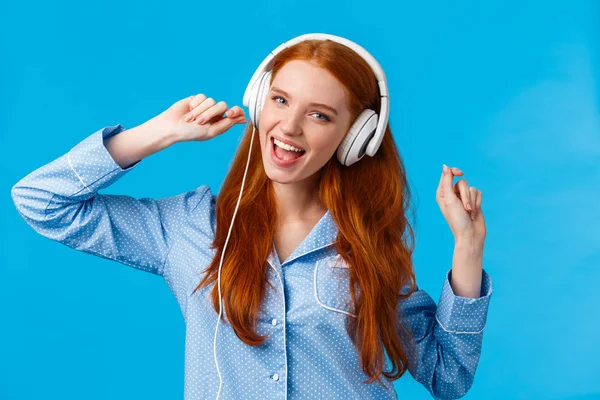 Menina acordar começando o dia com música. Joyful adolescente ruiva despreocupada dançando em roupas noturnas, levantando as mãos usando grandes fones de ouvido brancos cantando ao longo da música favorita, playlist, fundo azul — Fotografia de Stock