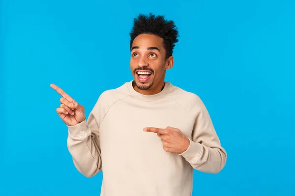 Dreamy inspired and amused, excited smiling african american man seeing first snow, looking at product, company banner, staring and pointing upper left corner fascinated, blue background — Stock Photo, Image