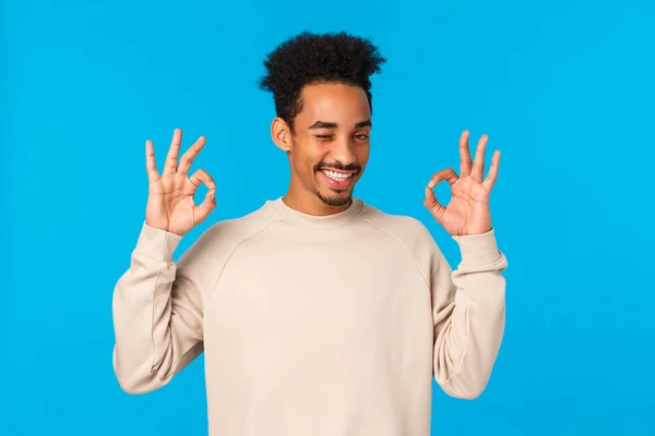 Cheeky and flirty, attractive sly african-american hipster guy checking out something awesome, showing okay, good or approval gesture, wink coquettish and smirk satisfied, blue background — Stock Photo, Image