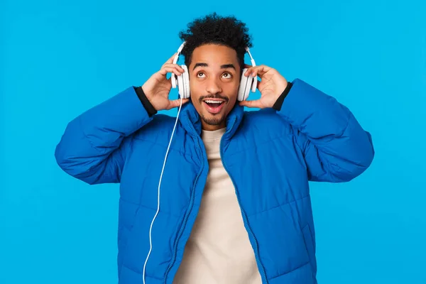 Chico disfrutando de buen sonido suave de los nuevos auriculares padres compraron para Navidad. Alegre hipster elegante hombre afroamericano en chaqueta de invierno acolchada, ponerse auriculares escuchar música sonriendo —  Fotos de Stock