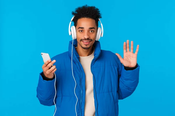 Hola, me alegro de verte. Alegre hombre afro-americano con estilo feliz en chaqueta de invierno acolchada, escuchar auriculares de música, mantenga la canción del interruptor del teléfono inteligente, saludando a la cámara, gesto de hola, fondo azul —  Fotos de Stock