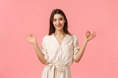 Sakin ol ve alışverişe gidelim. Güzel elbiseler giyen neşeli, küstah ve kadınsı genç esmer kadın, zen jesti gösteriyor, meditasyon yapıyor ve göz kırpıyor, pembe arka planda duruyor.