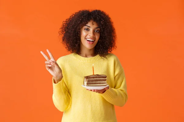 Tailliertes Porträt glückliche afrikanisch-amerikanische Frau in gelbem Pullover, zeigt Friedenszeichen und Käse, Geburtstagskind macht Foto mit Geburtstagstorte und Kerze, macht Wünsche, orangefarbener Hintergrund — Stockfoto