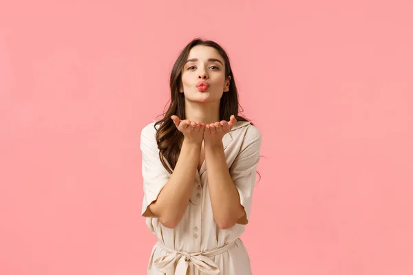 A mandar cumprimentos e cuidados. Encantador bonito e macio morena europeia com cabelo encaracolado, vestindo vestido de primavera, inclinando-se com as mãos perto de lábios dobrados, soprando beijo de ar com amor, de pé fundo rosa — Fotografia de Stock